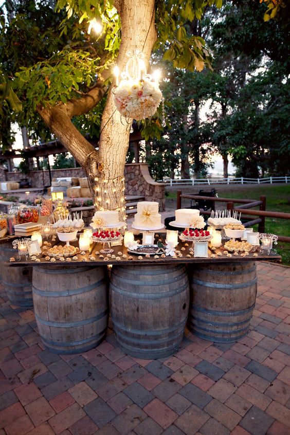 wesele w stylu rustykalnym - stół w plenerze w formie płyty ustawionej na trzech beczkach pod drzewem z podwieszonym pięknie udekorowanym żyrandolem